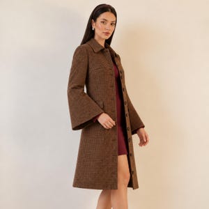 May include: A brown and black houndstooth patterned coat featuring a structured collar, long sleeves with flared cuffs, and a full-length button-down front closure. The coat is worn over a solid dark red dress, creating a layered appearance.