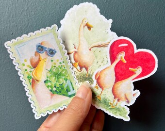 Może przedstawiać: Kolekcja fantazyjnych naklejek z ilustracjami kaczek. Jedna naklejka to projekt znaczka pocztowego z kaczką w okularach przeciwsłonecznych. Inne naklejki przedstawiają kaczki z czerwonym sercem i na trawiastym polu.