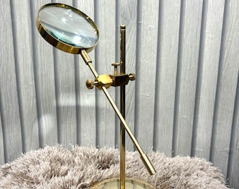 May include: A brass magnifying glass on a stand. The round lens is held by a brass frame and is adjustable. The stand has a round base and a vertical pole with an adjustable arm. The magnifying glass is designed for detailed viewing.