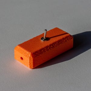 以下が含まれることがあります： 上部に銀色のトグルスイッチが付いたオレンジ色の長方形のオブジェクト。オブジェクトの端には小さな穴があります。このアイテムは灰色の表面に影を落としています。