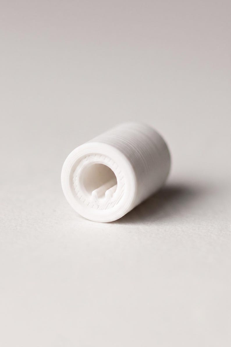 May include: A small, cylindrical white component with a hollow center and a visible internal notch. The exterior surface features fine, horizontal ridges consistent with 3D printing layers, creating a textured finish on the circular object.