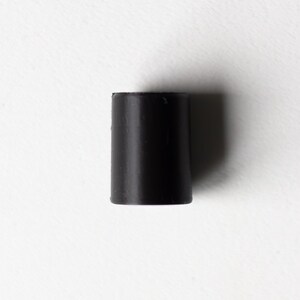 K&ouml;nnte beinhalten: Ein schwarzer, zylindrischer Gummistopfen, vor einem wei&szlig;en Hintergrund. Der Stopfen ist ein massives, opakes Schwarz mit einer glatten, schmucklosen Oberfl&auml;che. Das Objekt steht aufrecht und wirft einen dezenten Schatten.