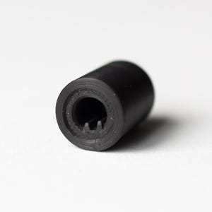 K&ouml;nnte beinhalten: Ein schwarzes zylindrisches Objekt mit einem ausgeh&ouml;hlten Ende und geriffelter Innenseite. Das Objekt befindet sich vor einem wei&szlig;en Hintergrund, wobei der Fokus auf seiner strukturierten Oberfl&auml;che und Form liegt. Das Design deutet auf eine mechanische oder industrielle Komponente hin.