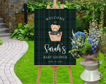 May include: A rectangular welcome sign featuring a dark green and navy blue plaid pattern. The sign displays a graphic of a teddy bear in a basket, with the text "WELCOME TO Sarah's BABY SHOWER APRIL 15, 2024" printed in white lettering.