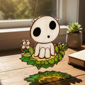 May include: A stained glass suncatcher featuring a white spirit character with large black eyes, suspended by a copper chain. The character is surrounded by green leaves and a flower, casting a colorful shadow on the wooden surface.