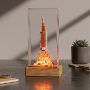 May include: A decorative rocket ship model inside a clear acrylic case, with a wooden base. The rocket is orange and white, with a fiery glow emanating from the base. A pen and coaster are in the foreground.