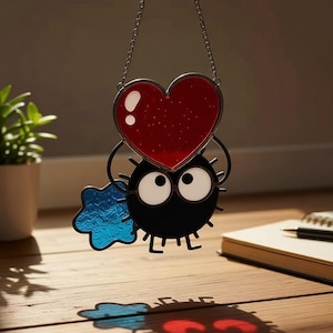 Könnte beinhalten: Ein Glasfenster-Suncatcher mit einer schwarzen Kreatur mit großen weißen Augen, die ein rotes Herz und einen blauen Stern hält. Der Suncatcher hängt an einer Kette und wirft einen Schatten auf eine Holzoberfläche. Im Hintergrund befindet sich eine kleine Pflanze in einem weißen Topf.
