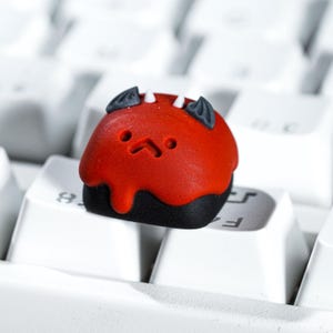 Puede incluir: Una tecla roja y negra con temática de diablo, con una cara de dibujos animados, cuernos y alas. La tecla está en un teclado blanco, añadiendo un toque único a cualquier configuración de teclado mecánico. Un accesorio divertido y peculiar.