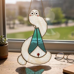 Könnte beinhalten: Ein skurriler Glasfenster-Sonnfänger in Form einer Cartoon-Figur. Der weiße Körper hat einen perlmuttartigen Glanz, mit einer türkis-grünen Dreiecksform in der Mitte. Ein kleiner roter Punkt befindet sich unten.