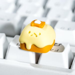 May include: A custom keycap shaped like a piece of toast with butter. The keycap is yellow and orange with a cute face and sits on a white keyboard. The butter is a square shape with a white dollop on top.