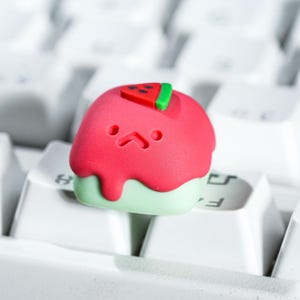 May include: A custom keycap in the shape of a watermelon slice, with a red top, green base, and a small watermelon slice on top. The keycap has a cute, cartoonish face and sits on a white keyboard.