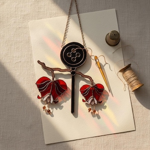 Puede incluir: Una pieza de arte en vidriera con un dise&ntilde;o de bot&oacute;n negro, un tallo y dos formas de flores rojas y blancas. Tambi&eacute;n hay un dedal, una aguja y un carrete de hilo, lo que sugiere un tema de costura o artesan&iacute;a.
