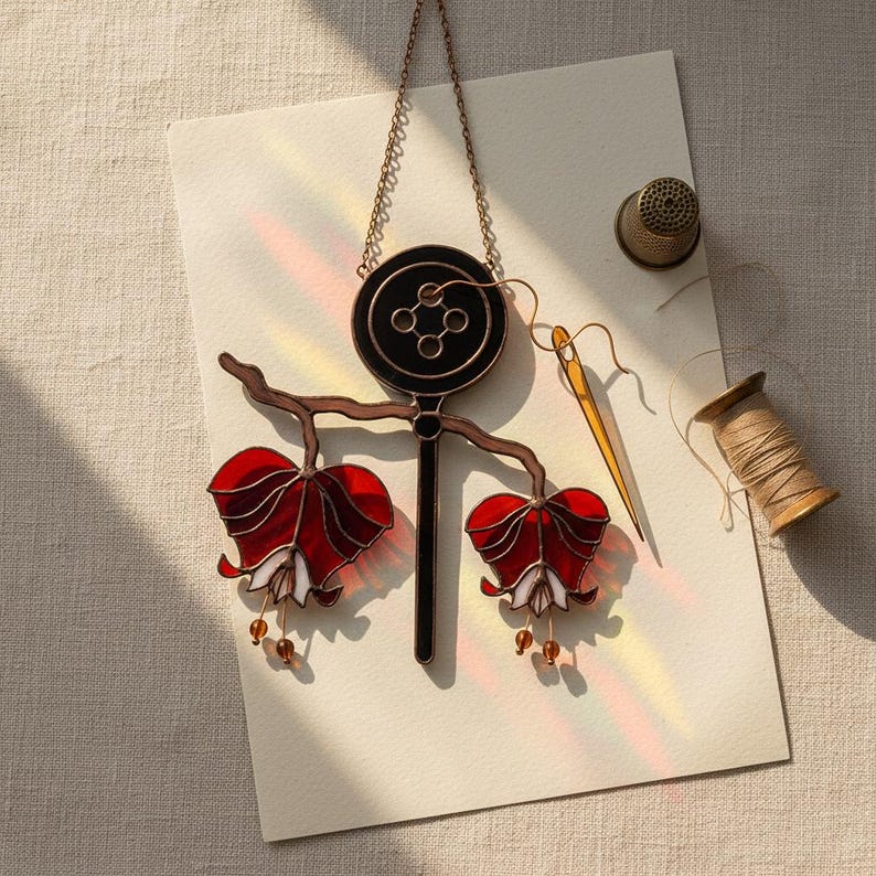 Puede incluir: Una pieza de arte en vidriera con un dise&ntilde;o de bot&oacute;n negro, un tallo y dos formas de flores rojas y blancas. Tambi&eacute;n hay un dedal, una aguja y un carrete de hilo, lo que sugiere un tema de costura o artesan&iacute;a.