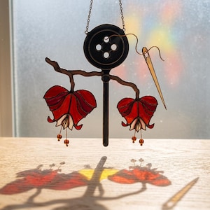 Puede incluir: Pieza de arte en vidriera que presenta un bot&oacute;n negro con cuatro agujeros, una aguja y dos formas de flores rojas y blancas. La pieza est&aacute; suspendida por una cadena, proyectando una sombra sobre una superficie de madera. Las flores tienen peque&ntilde;as cuentas colgantes.