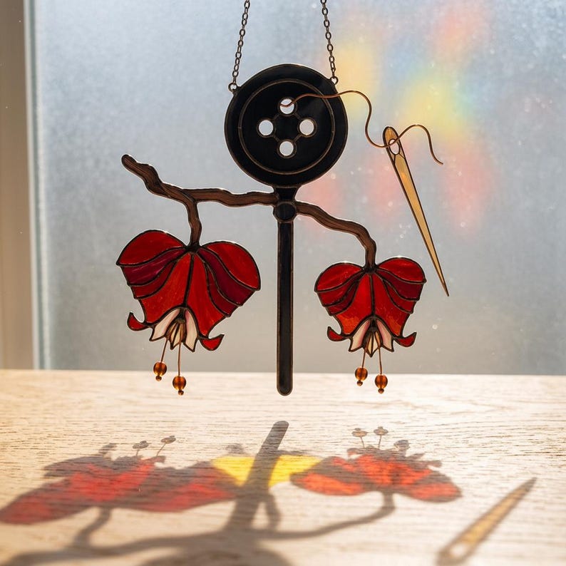 Puede incluir: Pieza de arte en vidriera que presenta un bot&oacute;n negro con cuatro agujeros, una aguja y dos formas de flores rojas y blancas. La pieza est&aacute; suspendida por una cadena, proyectando una sombra sobre una superficie de madera. Las flores tienen peque&ntilde;as cuentas colgantes.