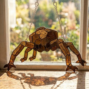 May include: A stained glass spider suncatcher with a geometric body and articulated legs. The spider is suspended by a silver chain. The glass is a mix of amber and brown tones, creating a warm, inviting glow. The spider's shadow is cast on the wooden surface below.