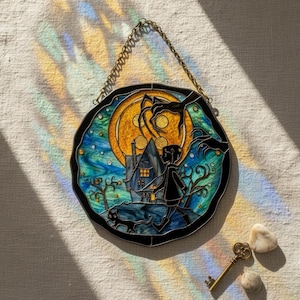 Puede incluir: Panel de vidriera que representa una escena caprichosa con un ni&ntilde;o, un gato y una casa bajo la luna llena. La obra de arte utiliza tonos de azul, amarillo y negro, con una cadena de metal decorativa para colgar. Una llave antigua y piedras est&aacute;n debajo.