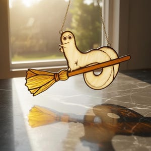 May include: A whimsical stained glass suncatcher featuring a cartoonish creature riding a broom. The creature is cream-colored with black outlines, and the broom is brown with yellow bristles. The suncatcher hangs from a delicate chain, perfect for a window display.