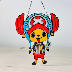 May include: A colorful stained glass decoration featuring a cartoon character with a yellow face, red hat, and blue accents. The character has a cheerful expression and is suspended by a chain. The design is outlined in black.