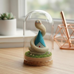 Puede incluir: Una escultura caprichosa de una serpiente enrollada alrededor de una pequeña casa con techo azul, dentro de una cúpula de cristal. La base es de corcho y césped artificial. La serpiente es de color beige claro y la casa tiene un punto rojo.
