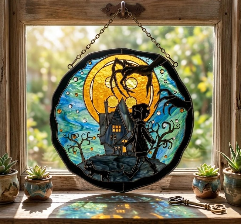 Puede incluir: Vidriera que representa una escena espeluznante con un ni&ntilde;o, una casa y un gato silueteados contra una luna llena. La obra de arte est&aacute; enmarcada con un borde negro y cuelga de una cadena. El fondo tiene tonos azules y amarillos.