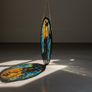 Puede incluir: Un atrapasoles ovalado de vidriera con borde negro, colgado de una cadena dorada. La obra de arte presenta una escena vibrante con tonos naranja, azul y verde, proyectando una sombra colorida sobre la superficie inferior.