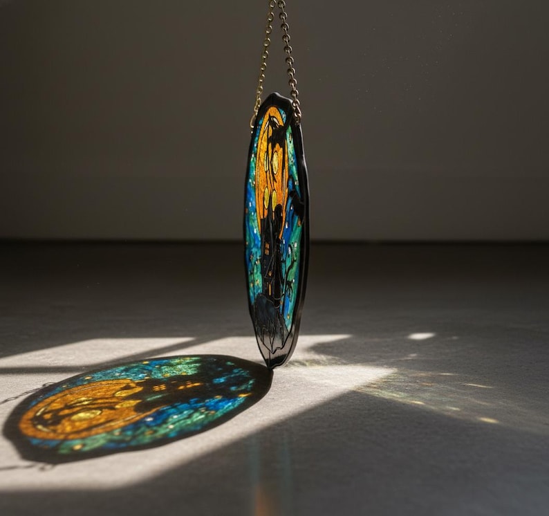 Puede incluir: Un atrapasoles ovalado de vidriera con borde negro, colgado de una cadena dorada. La obra de arte presenta una escena vibrante con tonos naranja, azul y verde, proyectando una sombra colorida sobre la superficie inferior.