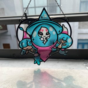May include: A stained glass suncatcher featuring a whimsical cartoon character with a blue and pink color scheme. The design includes a witch hat, a skull-like face, and two smaller characters. The suncatcher is framed by a black metal outline and hangs from a chain.