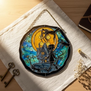 Puede incluir: Panel de vidriera redondo que representa una escena espeluznante con una casa, una figura y un gato bajo la luna llena. La obra de arte utiliza tonos de azul, amarillo y negro, con una cadena decorativa para colgar. Se ven dos llaves antiguas.