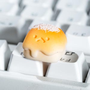 May include: A custom artisan keycap in the shape of a cartoon character, featuring a yellow and orange color scheme with a white base and a sprinkle-like texture on top. The keycap is resting on a white keyboard.