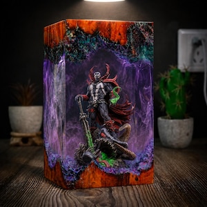 Puede incluir: Una pieza de arte de resina rectangular con una figura de acción de Spawn. La figura está sobre un monstruo, sosteniendo una espada, dentro de un bloque de resina con tonos naranja, morado y verde. La pieza está iluminada desde arriba.