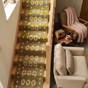 Könnte beinhalten: Ein langer, rechteckiger olivgrüner Treppenläufer mit einem sich wiederholenden geometrischen Muster in Weiß, Gelb und Braun. Der Teppich hat eine Fransenkante am unteren Ende und ist vertikal über eine Holztreppe ausgelegt, wobei er mehrere Stufen bedeckt.
