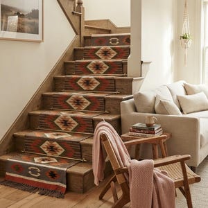 Könnte beinhalten: Eine Holztreppe mit einem gewebten Treppenläufer mit geometrischem Muster in Braun-, Rot- und Cremetönen, befestigt durch Messingstangen. Der Läufer hat eine gefranste Kante an der untersten Stufe und zeigt wiederkehrende Rautenmotive.