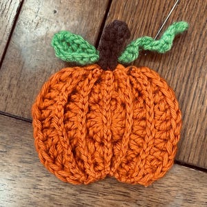 Puede incluir: Una calabaza hecha a mano con ganchillo en hilo naranja, con un tallo marrón y hojas verdes. La calabaza es redonda con costillas verticales, ideal para la decoración de otoño. Una aguja se inserta cerca de las hojas.