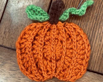 May include: A handmade crochet pumpkin in orange yarn, featuring a brown stem and green leaves. The pumpkin is round with vertical ribbing, perfect for fall decor. A needle is inserted near the leaves.