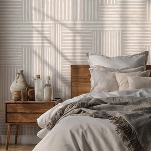 Op de afbeelding: Behang met een herhalend geometrisch patroon van afwisselende verticale en horizontale witte strepen op een lichtbeige achtergrond. Het ontwerp creëert een rasterachtig effect over het wandoppervlak en zorgt voor een getextureerd uiterlijk.