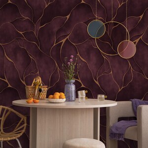 Peut inclure: Un papier peint bordeaux profond orné d'un motif abstrait de lignes dorées fluides. Le mur est décoré de trois suspensions circulaires bleu, or et marron. Une table ronde en bois clair est placée devant, avec un vase de fleurs et des oranges.