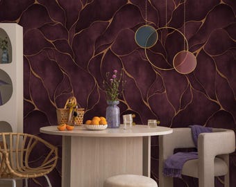 Op de afbeelding: Een diepbordeauxrood behang met een vloeiend, abstract goudkleurig lijnpatroon. De muur is versierd met drie cirkelvormige wandhangers in blauw, goud en kastanjebruin. Een ronde tafel van licht hout staat ervoor, met een vaas bloemen en sinaasappels.