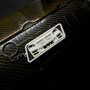 May include: A white keychain featuring a car's front grill design. The keychain is attached to a silver metal ring and chain. The background has a carbon fiber pattern.