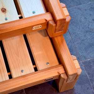 Op de afbeelding: Close-up van een houten bedframe met een natuurlijke afwerking. Het frame is gemaakt van lichtgekleurde houten planken en heeft stevige, in elkaar grijpende hoekverbindingen. Metalen schroeven bevestigen de componenten van het frame. Het bedframe is ontworpen voor duurzaamheid en een natuurlijke uitstraling.