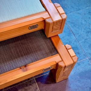 Può includere: Lettino per animali domestici in legno a due livelli con finitura marrone chiaro. Il livello superiore ha una superficie a rete blu chiaro, mentre il livello inferiore ha una rete marrone scuro. Gli angoli presentano un design scolpito unico. Piccole targhette metalliche sono visibili.