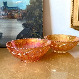 May include: Two iridescent orange glass bowls with star and geometric patterns. The bowls have a scalloped rim and sit on a table. A painting of a landscape and a gold framed mirror are in the background.