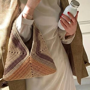 Puede incluir: Bolso de crochet hecho a mano en tonos beige, morado y naranja. El bolso tiene forma triangular con un patrón de rayas. Una botella blanca con tapa plateada se sostiene en el fondo.