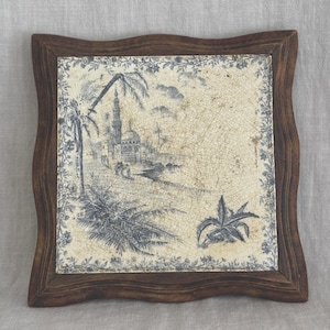 May include: A square ceramic tile with a crackled cream finish and blue ink illustration of a coastal landscape featuring a mosque, palm trees, and foliage. The tile is framed in a dark brown wooden border with a wavy, scalloped edge design.