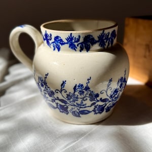 Może przedstawiać: Kremowy ceramiczny dzban z zaokrąglonym korpusem i uchwytem. Dzban jest ozdobiony niebieskimi wzorami kwiatowymi wokół krawędzi i korpusu. Projekt przedstawia stylizowane kwiaty i winorośl, tworząc klasyczną, vintage estetykę.