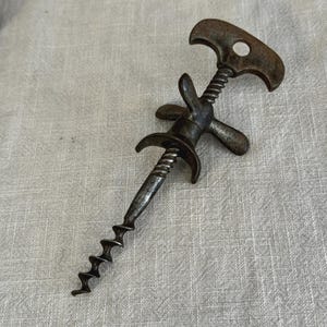 Peut inclure: Un tire-bouchon en métal vintage présentant une patine sombre et usée. L'outil comprend une poignée en forme de T avec un trou circulaire, une tige filetée centrale et une paire de bras latéraux incurvés s'étendant au-dessus de la mèche.