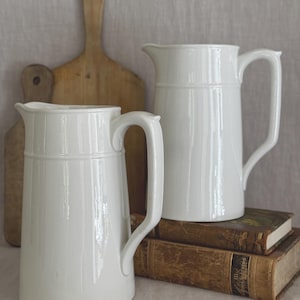Può includere: Due brocche in ceramica bianca con finiture lisce e lucide, manici curvi e beccucci svasati. Le brocche presentano un design semplice a corpo cilindrico con un sottile dettaglio a rilievo orizzontale vicino al bordo superiore.