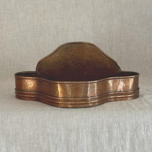 May include: A wall-mounted copper planter or storage vessel featuring a curved, raised back panel and a wide, shallow basin with a hammered texture. The metal has a warm, aged patina with visible variations in tone across the entire surface.