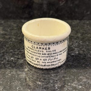 Può includere: Un piccolo barattolo di ceramica bianco sporco con un bordo arrotondato e un'etichetta avvolta attorno al corpo. L'etichetta presenta un testo nero che recita "CLARKES MIRACULOUS SALVE" e altri dettagli. Il barattolo è probabilmente un oggetto antico o vintage.