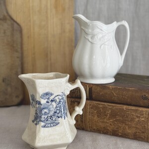 May include: A cream-coloured ceramic pitcher with a blue floral transfer pattern and a matching white ceramic pitcher featuring a raised leaf design. Both pitchers have a glossy finish with visible fine crackling across their surfaces.